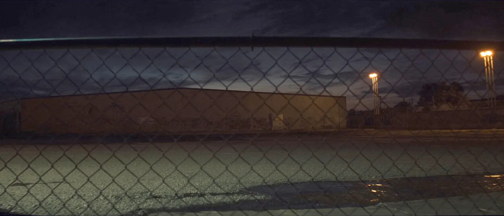 A chain link fence at night with floodlit ground and a graffiti wall beyond