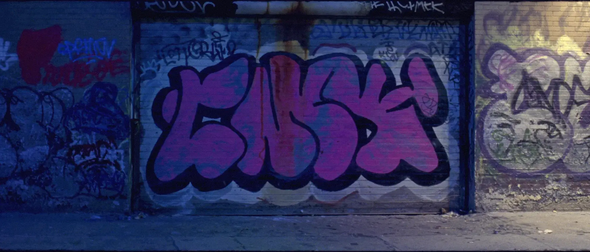 A steel shutter and brick wall layered with graffiti tags and one violet piece