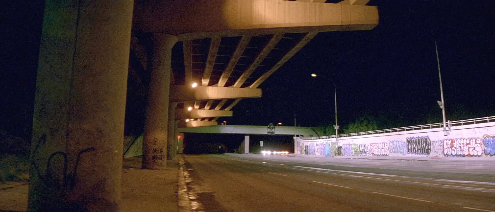 A freeway underpass at night, graffiti along the retaining wall under sodium light
